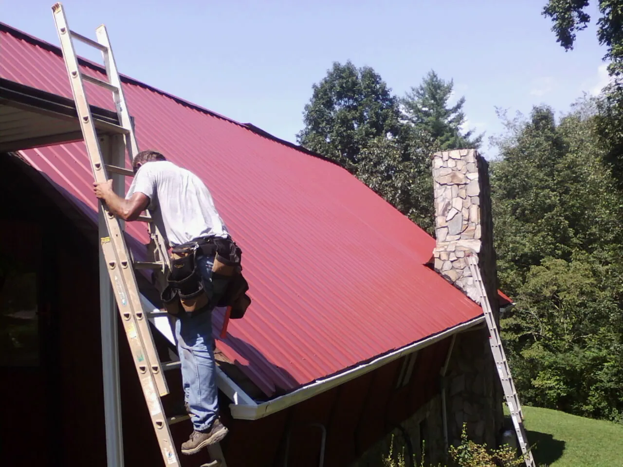 Roofer on a ladder beside a red metal roof and stone chimney — Soffit & Fascia Repair by Roofing Brothers in Fishersville